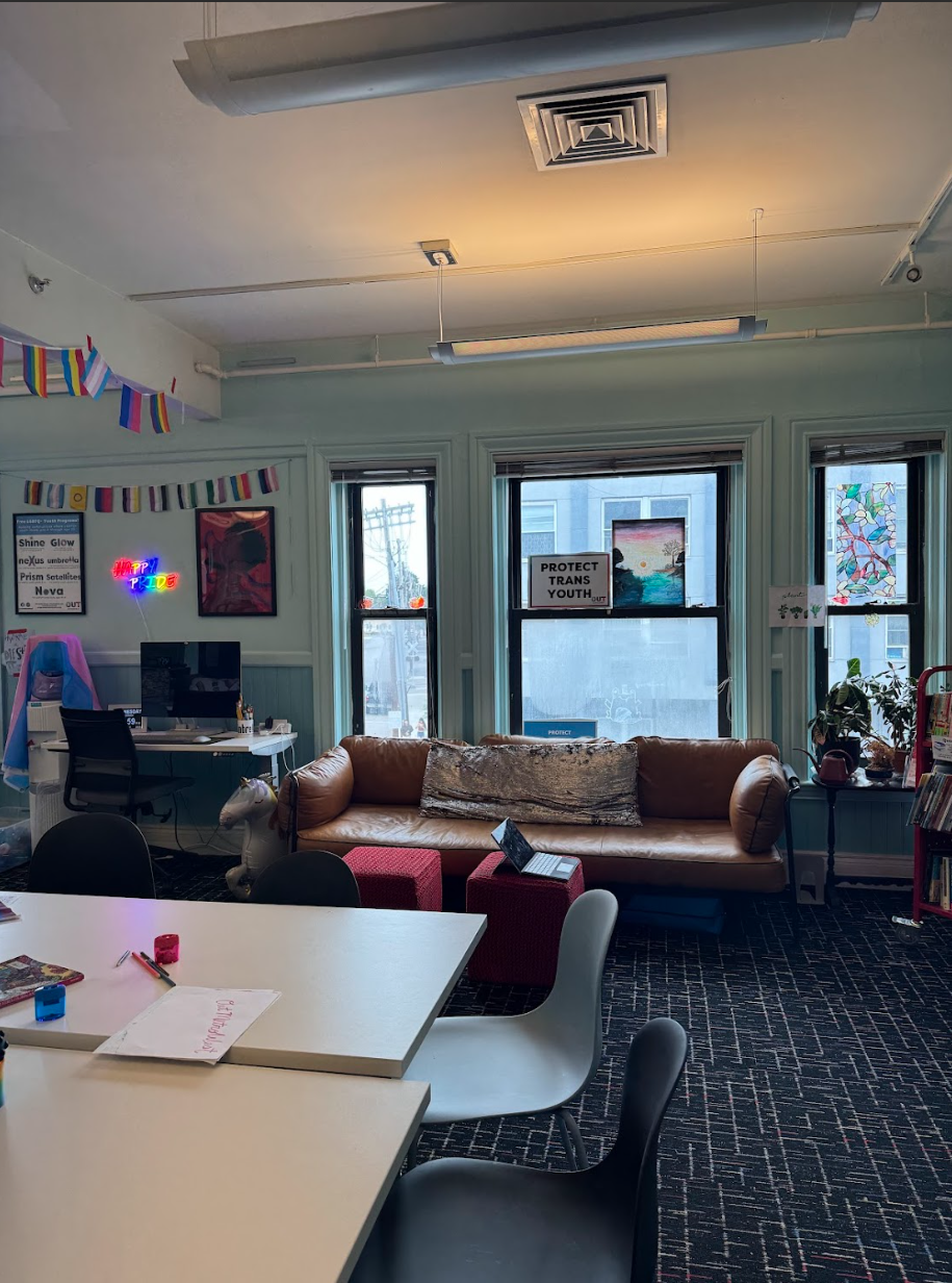 Cozy room with a brown leather couch, windows, and tables. Pride flag banner and desk visible.
