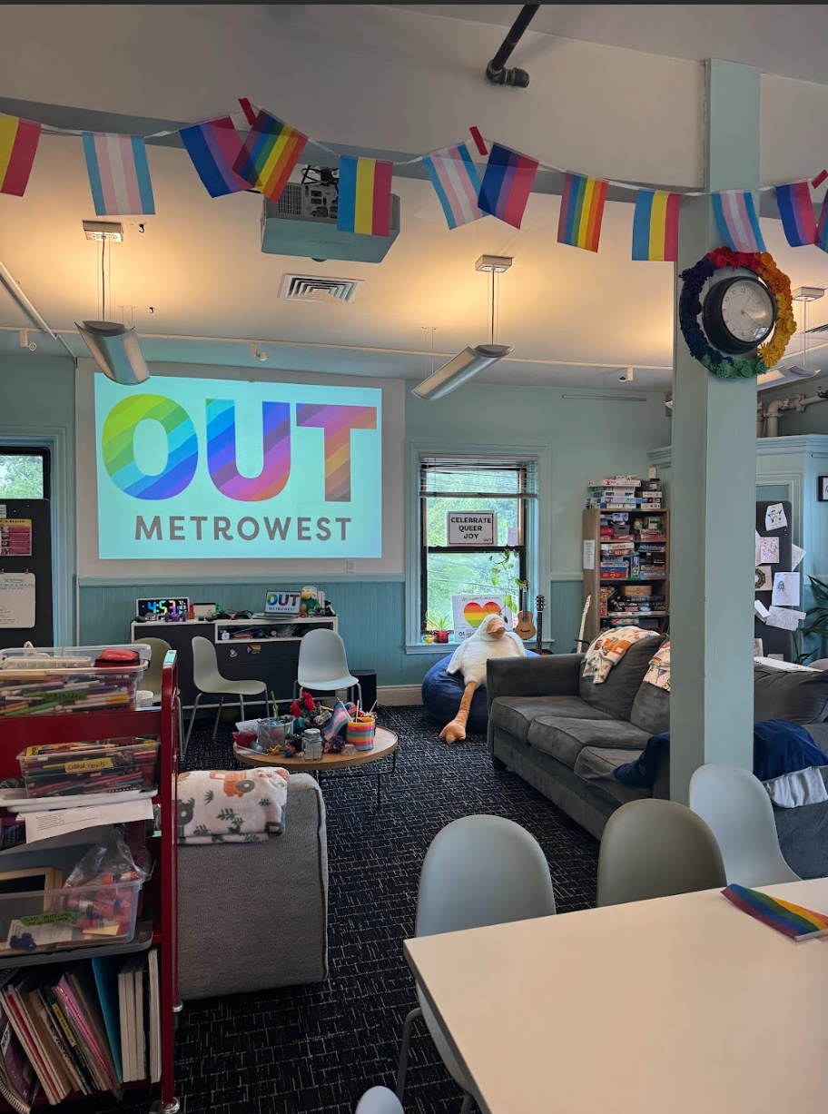 Interior room with "OUT METROWEST" projected on screen, adorned with Pride flags and a person on the floor.