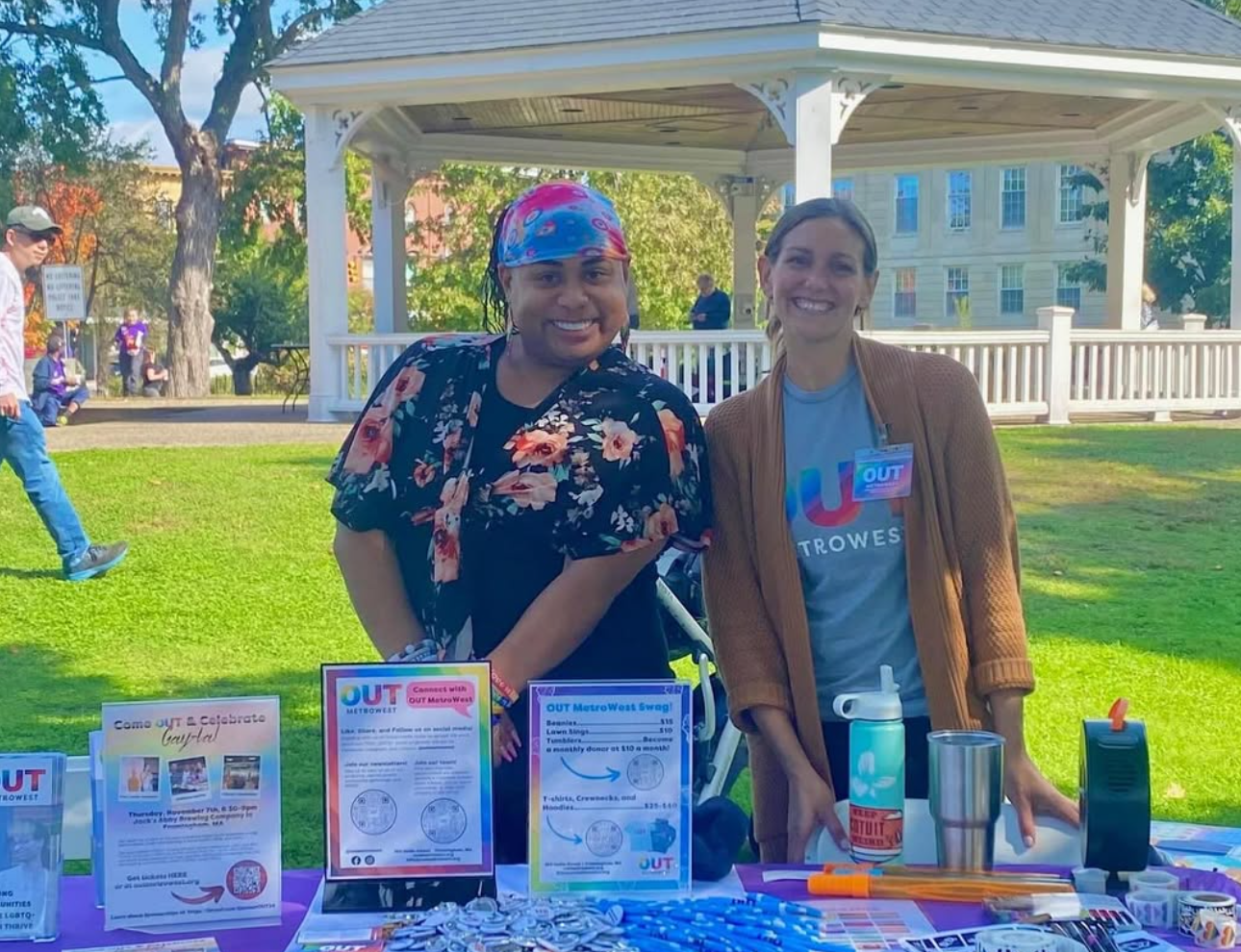 Two people at a table in a park, smiling. Gazebo and green grass in the background.