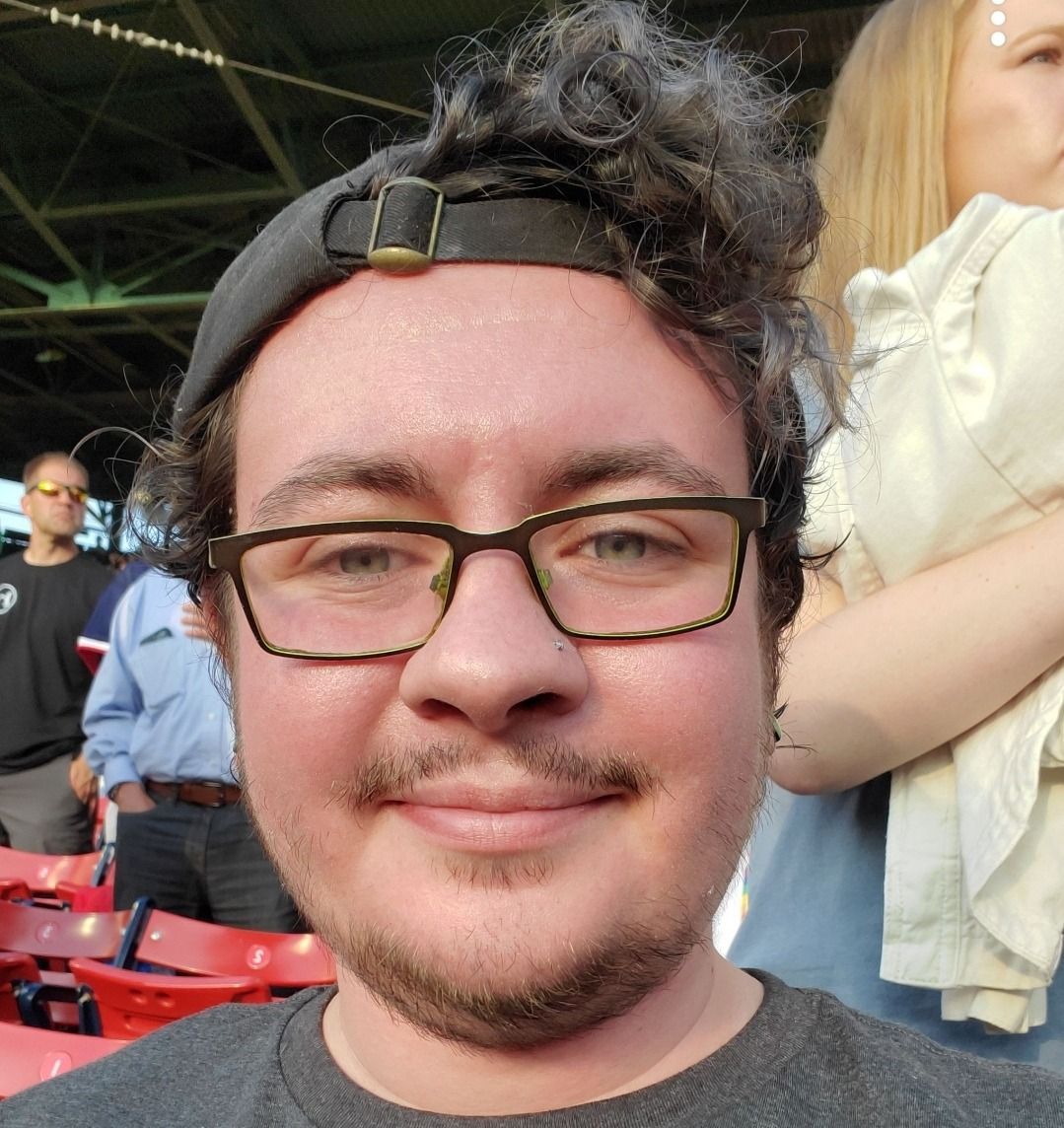 Person with glasses and a cap, smiling at a baseball game, with a crowd and red seats in the background.