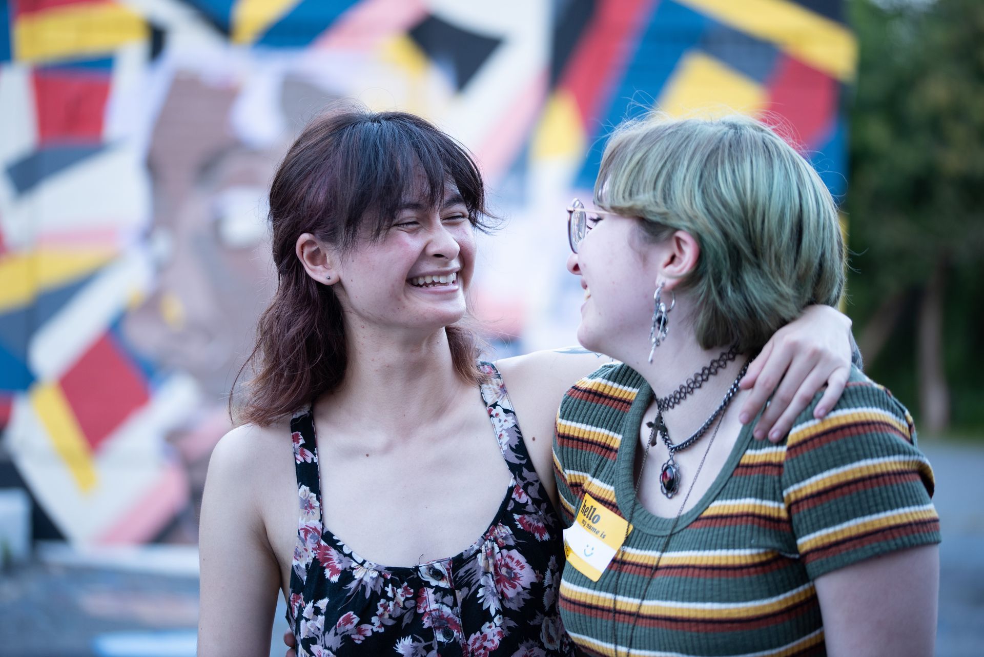Two people smiling, arm around each other. One with green hair, striped shirt, other with floral top.