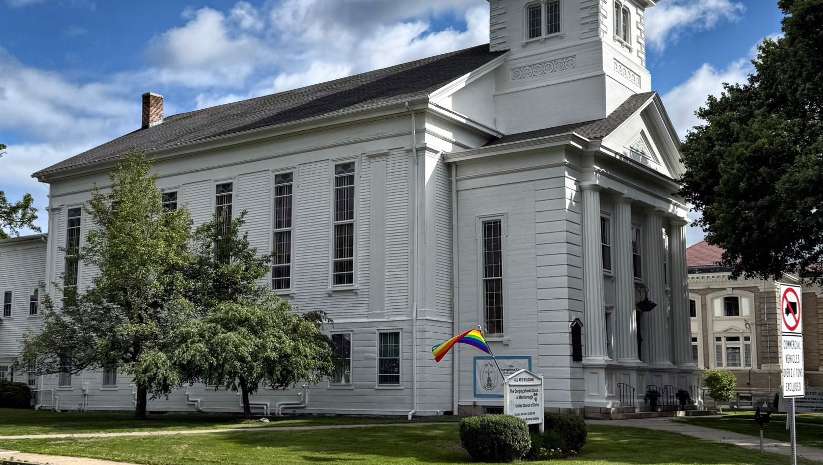 Exterior of the Westborough Church, with rainbow flag and sign; green grass and trees in front.