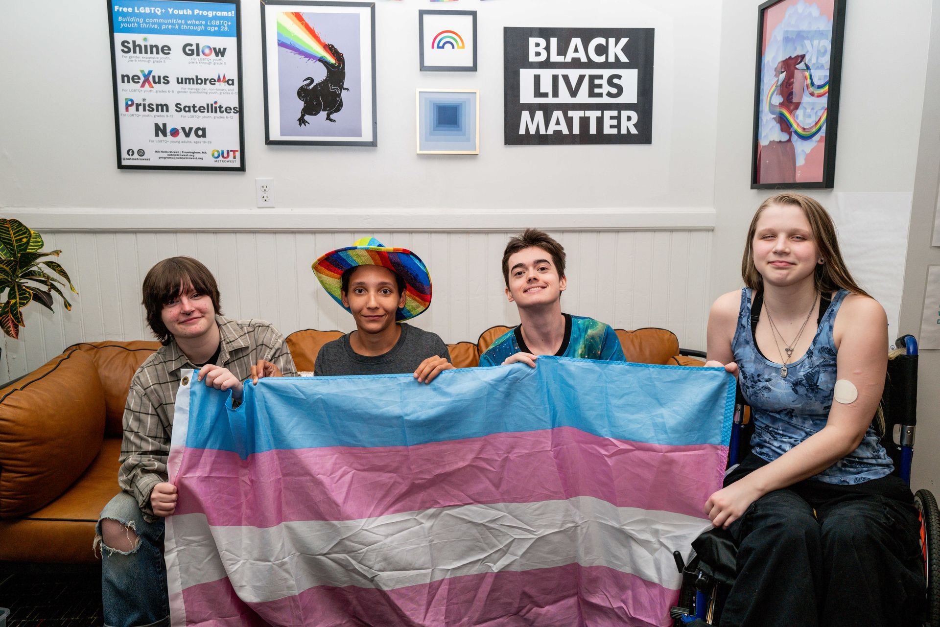 Four people holding a trans pride flag, sitting on a couch. Background: art, a 