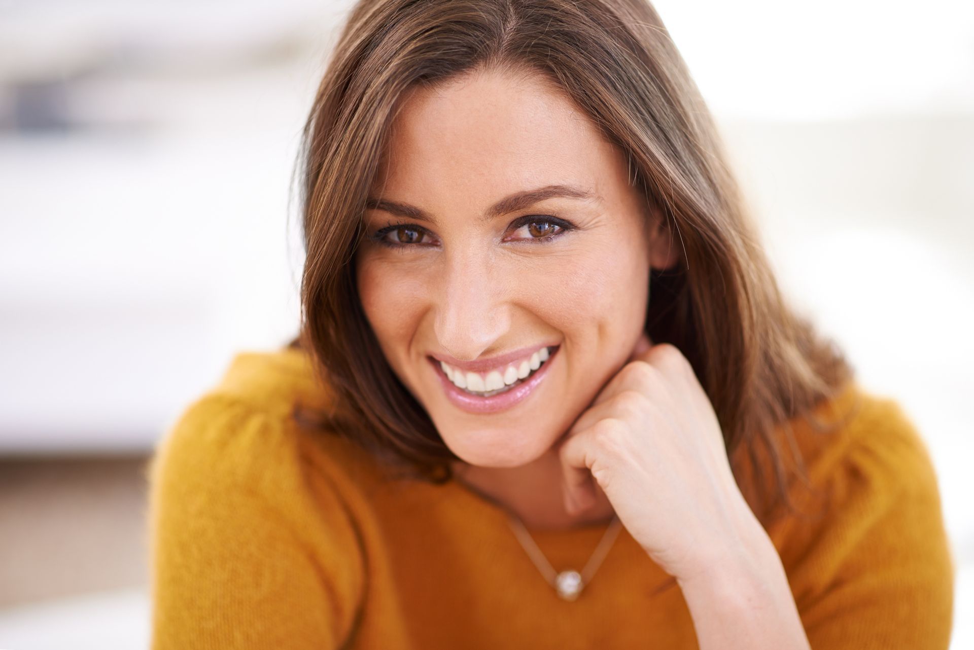 Woman with brown hair, smiling, rests chin on hand, wearing a yellow sweater.