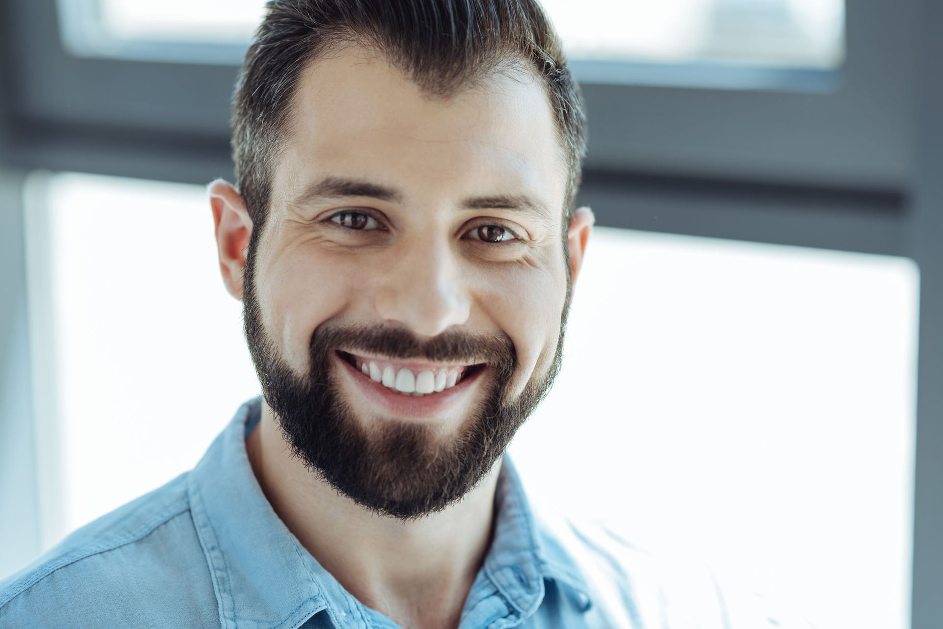 Lachende man met donker haar en een baard, gekleed in een blauw shirt, in een felverlichte ruimte.