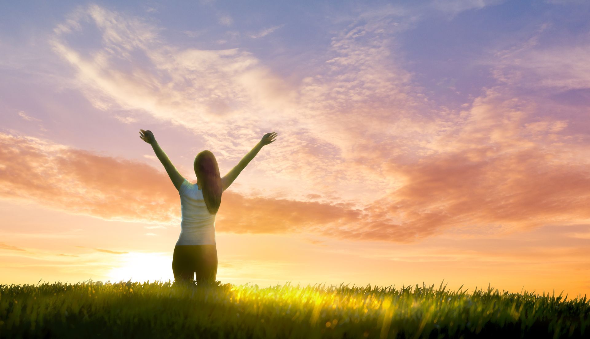 Woman with arms raised, silhouetted against a colorful sunset over a grassy field, expressing joy.