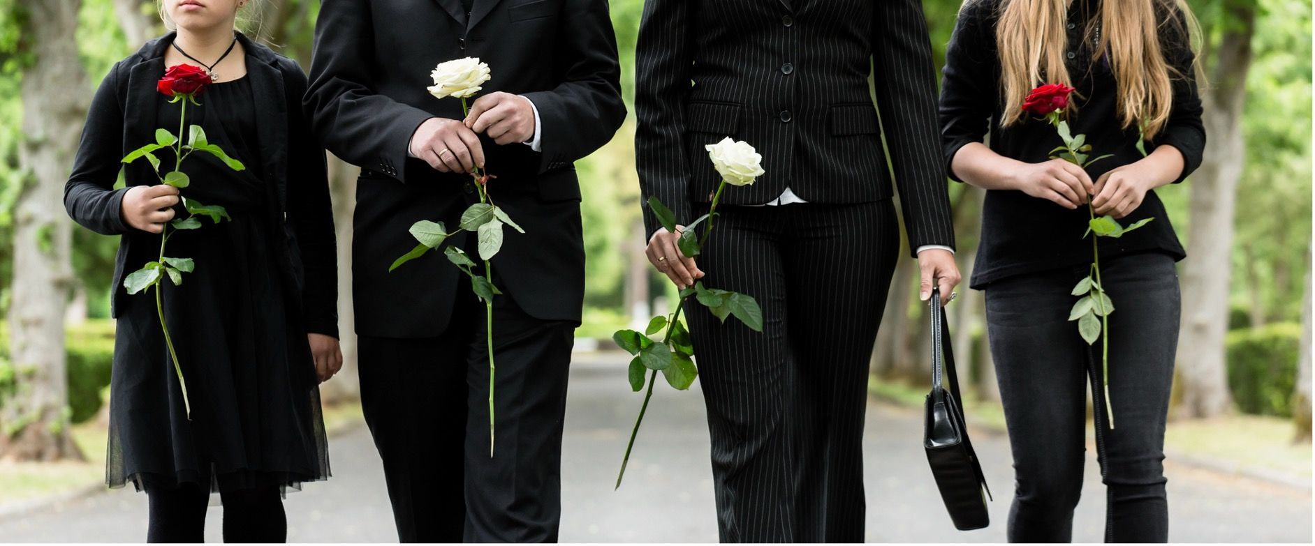 Four people in black clothing hold roses, walking down a tree-lined path.