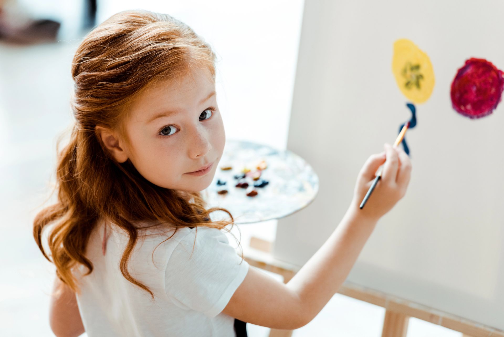 Young person with reddish hair painting on a canvas, holding a palette of paints.
