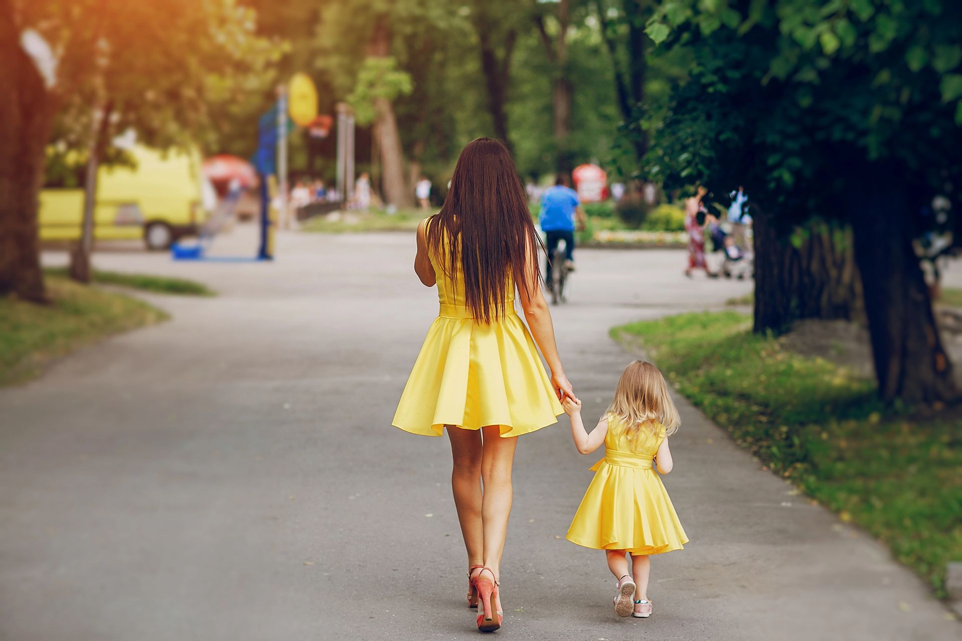 Woman and child in matching yellow dresses hold hands walking down a path in a park.