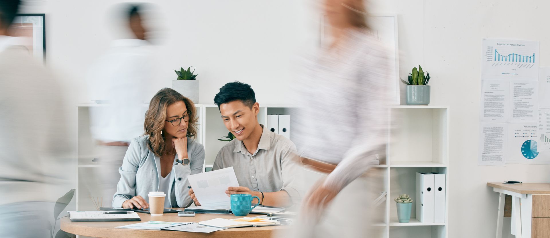 Two people at a desk review paperwork as blurred figures move through a bright office.