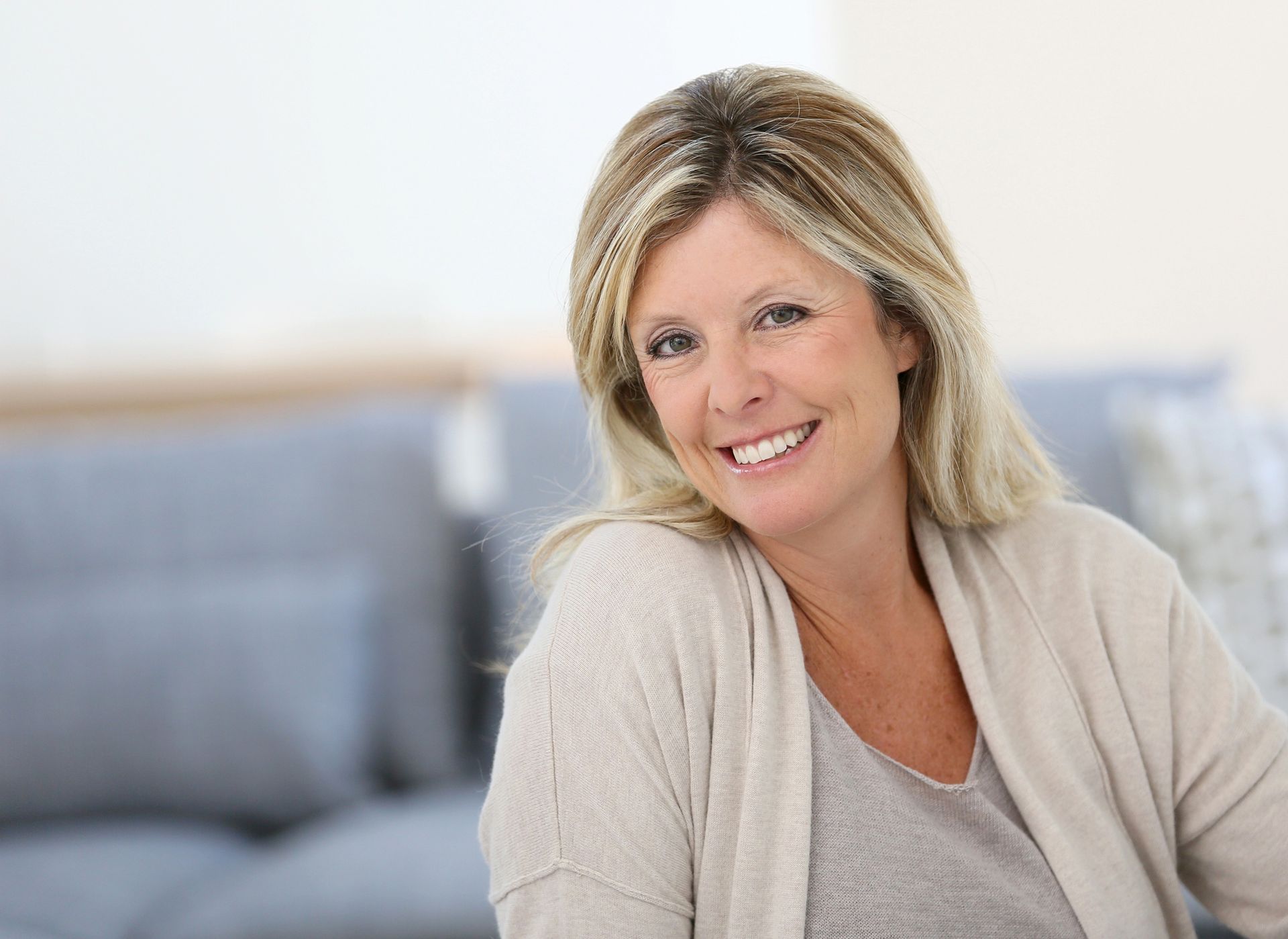 Woman with blonde hair smiling, wearing a light cardigan, sitting in a living room with gray couches.