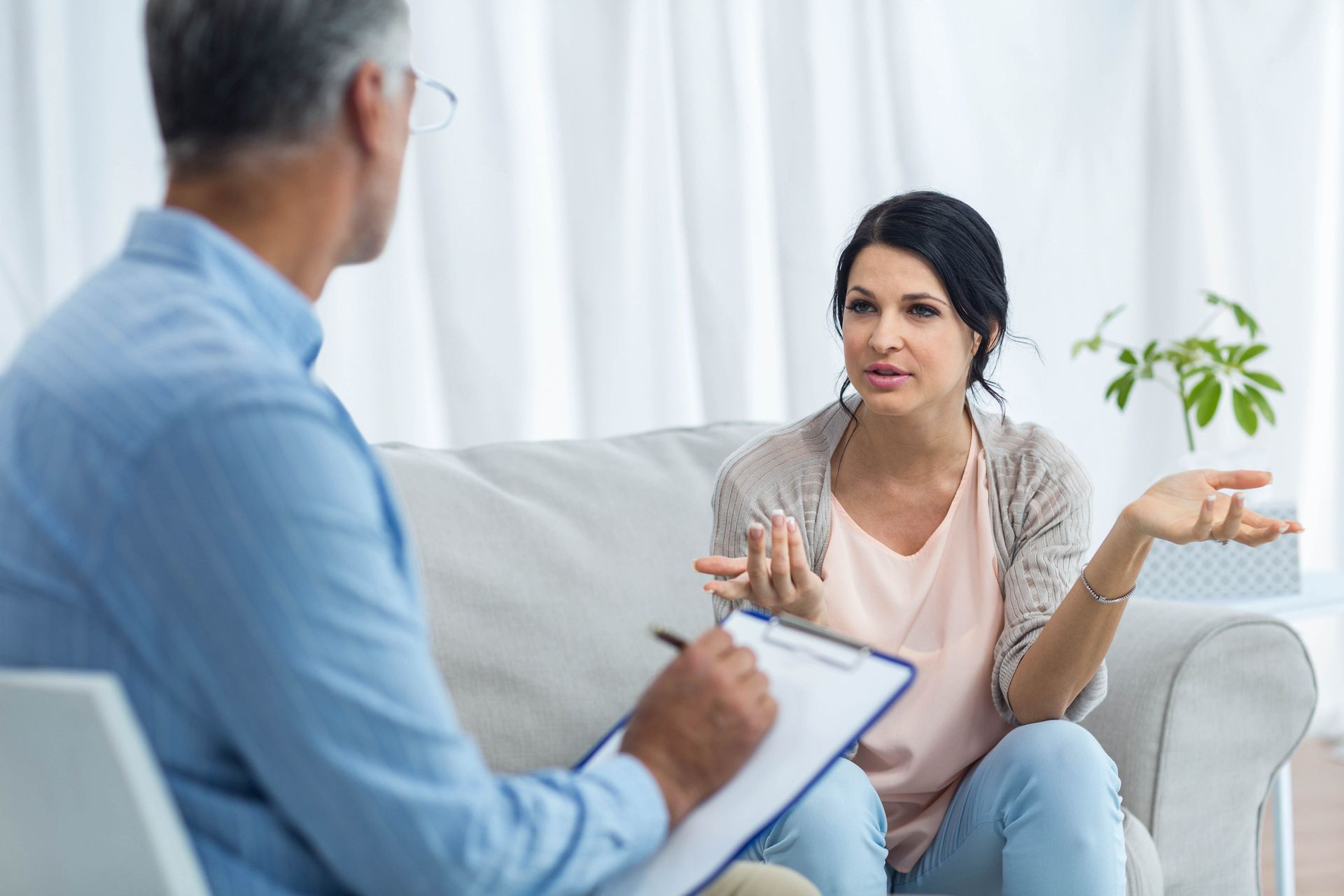 Woman talking with professional coach, gesturing, in a counseling setting. Professional coach takes notes on a clipboard.