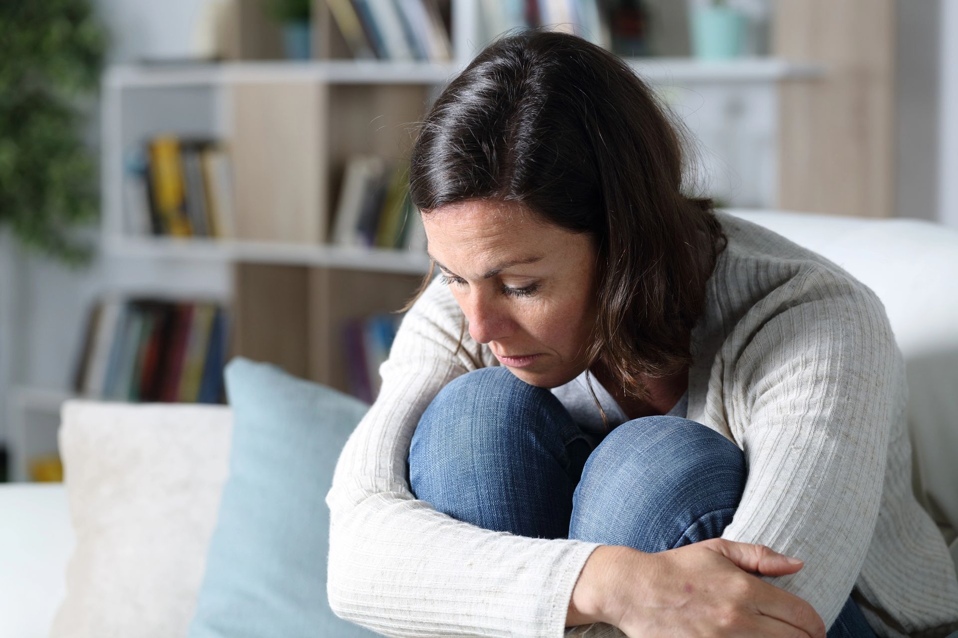 Woman in a sweater and jeans sits curled up on a couch, looking down, appearing sad.