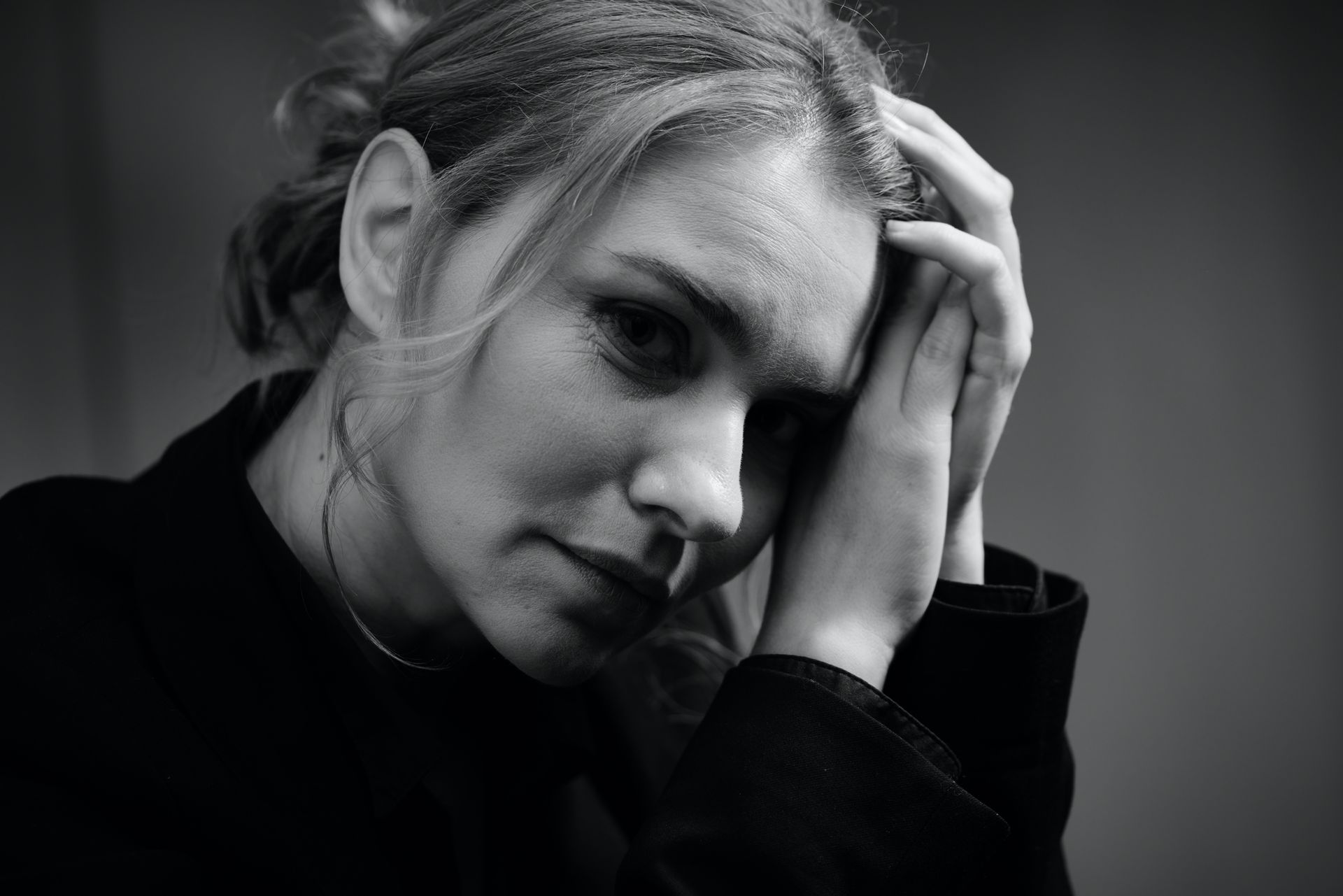 Woman with hands clasped near her face, gazing downward. Black and white photo, neutral background.