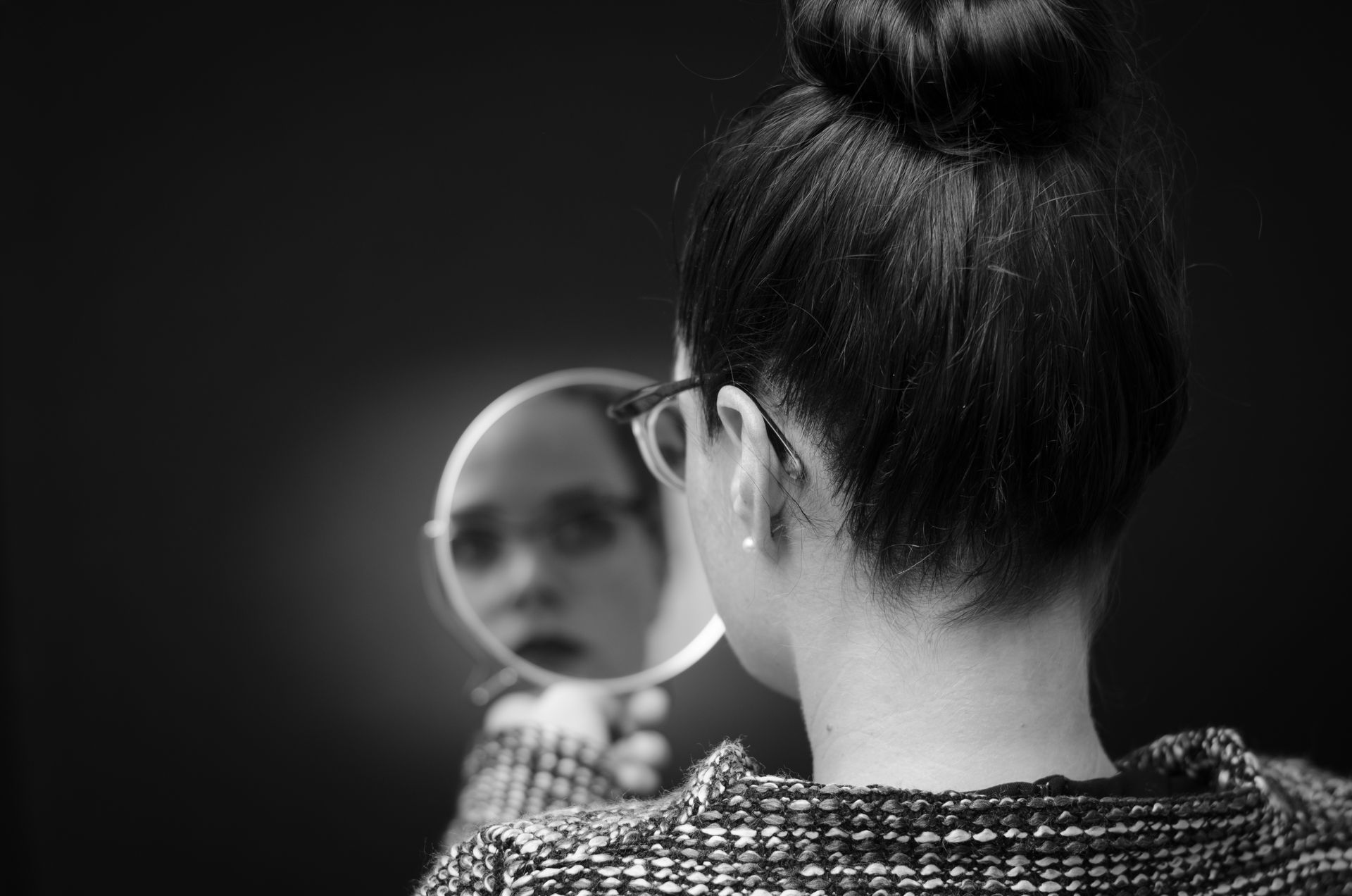 Woman looks in a hand mirror, dark background.
