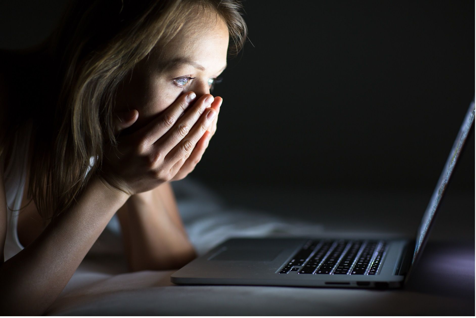Woman in dark room, looking at laptop with concerned expression, hands covering mouth.