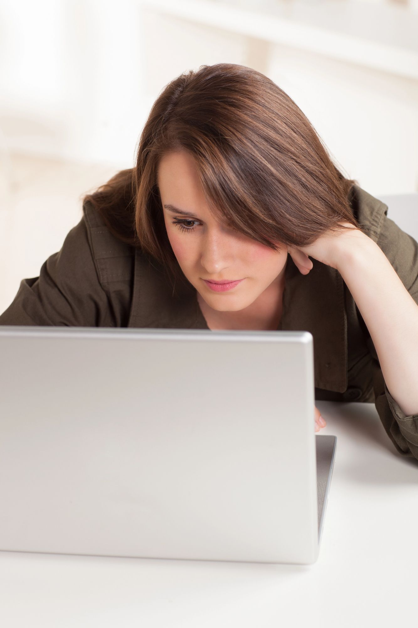 Woman in olive blazer looks intently at laptop on a white table.