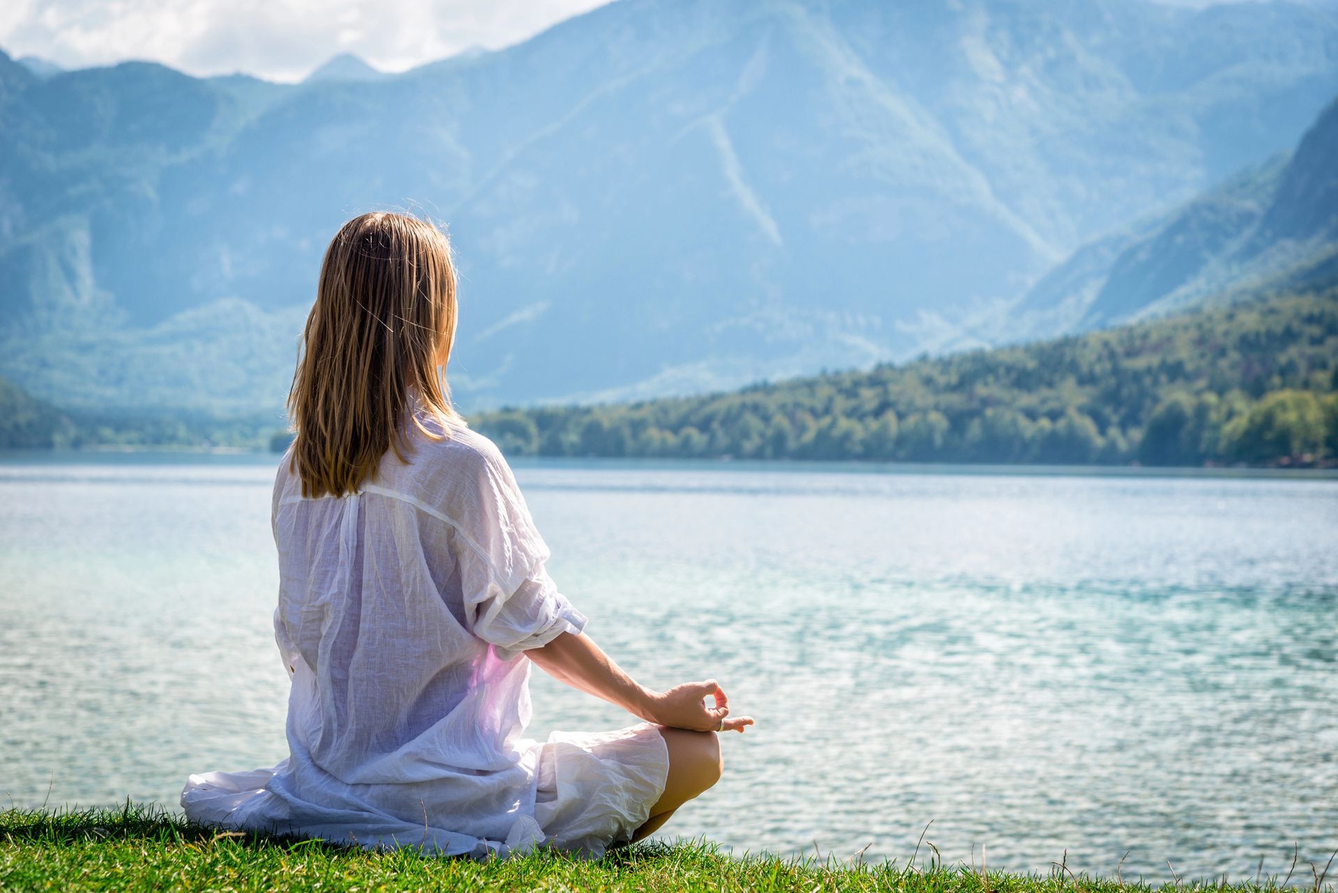 Woman meditating near lake, mountains in background. She is wearing white, outdoors.