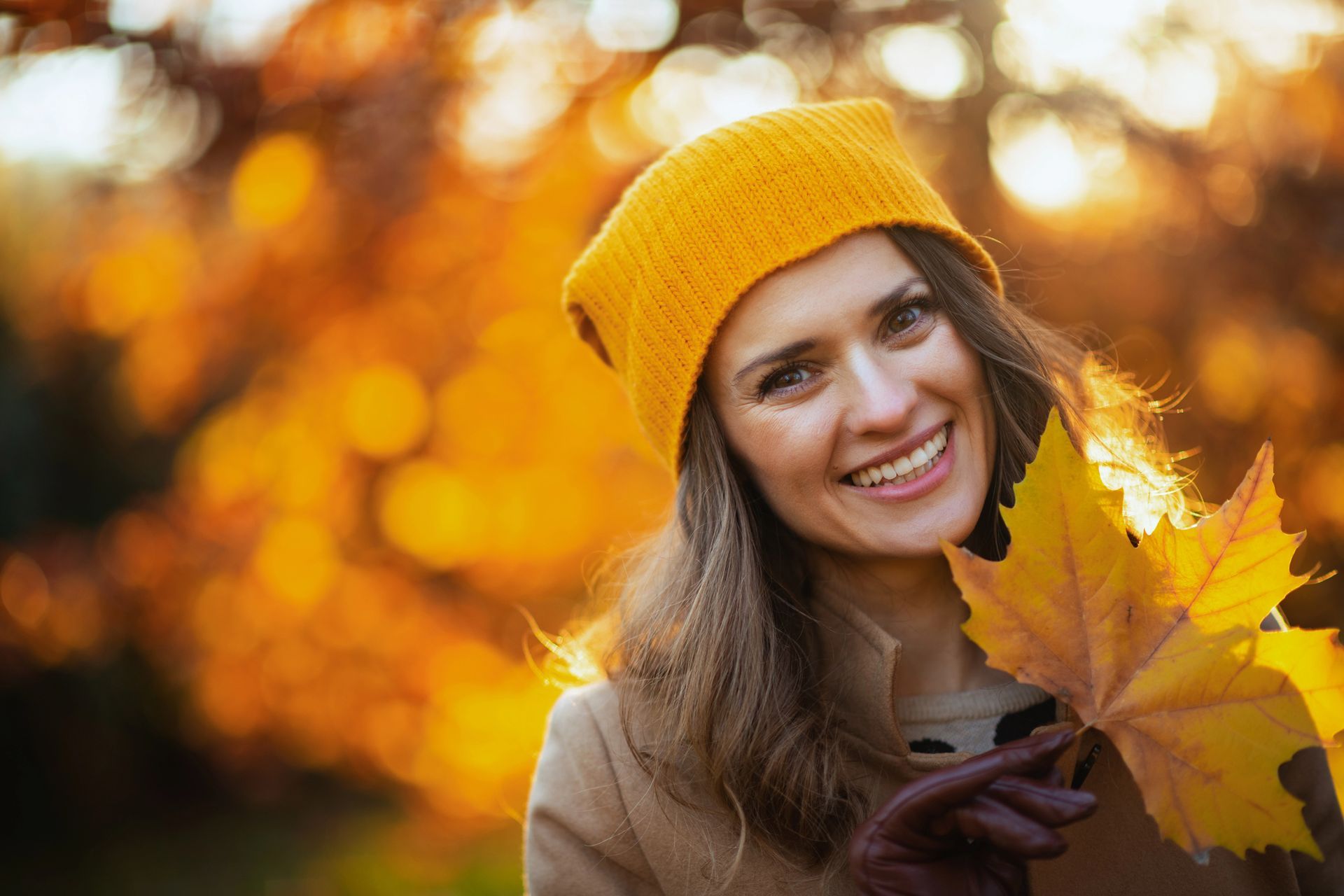 Woman in yellow hat smiles, holding yellow maple leaf in front of autumn foliage.