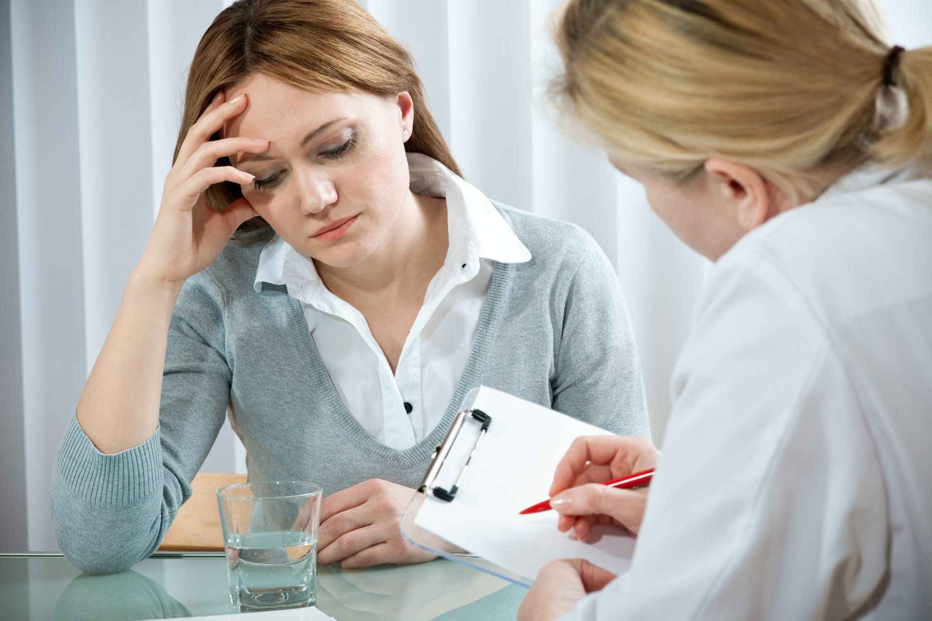 Woman with a pained expression sits at a desk, consulting with a healthcare professional who is writing on a clipboard.