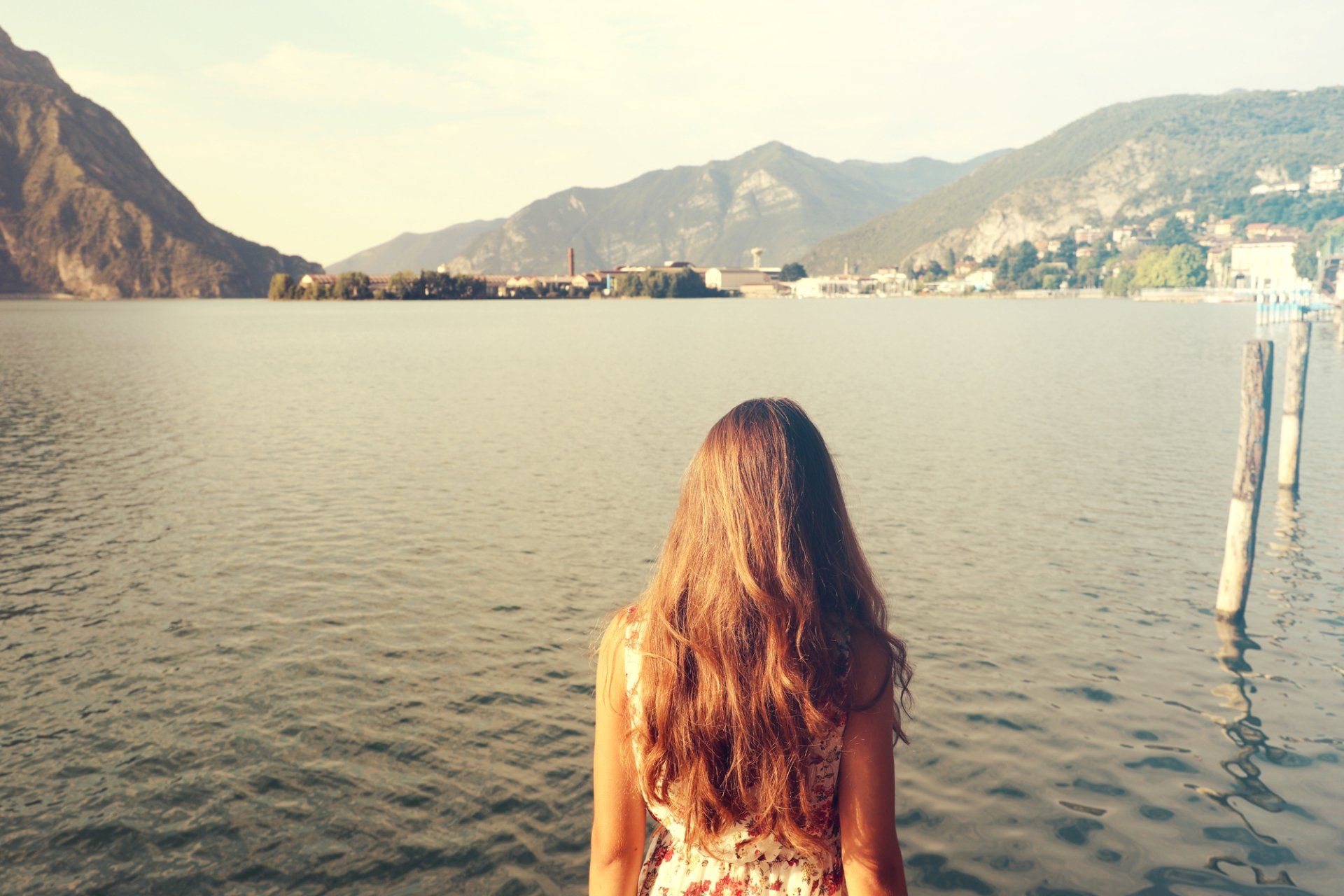 Woman with long brown hair gazes at a lake surrounded by mountains on a sunny day.