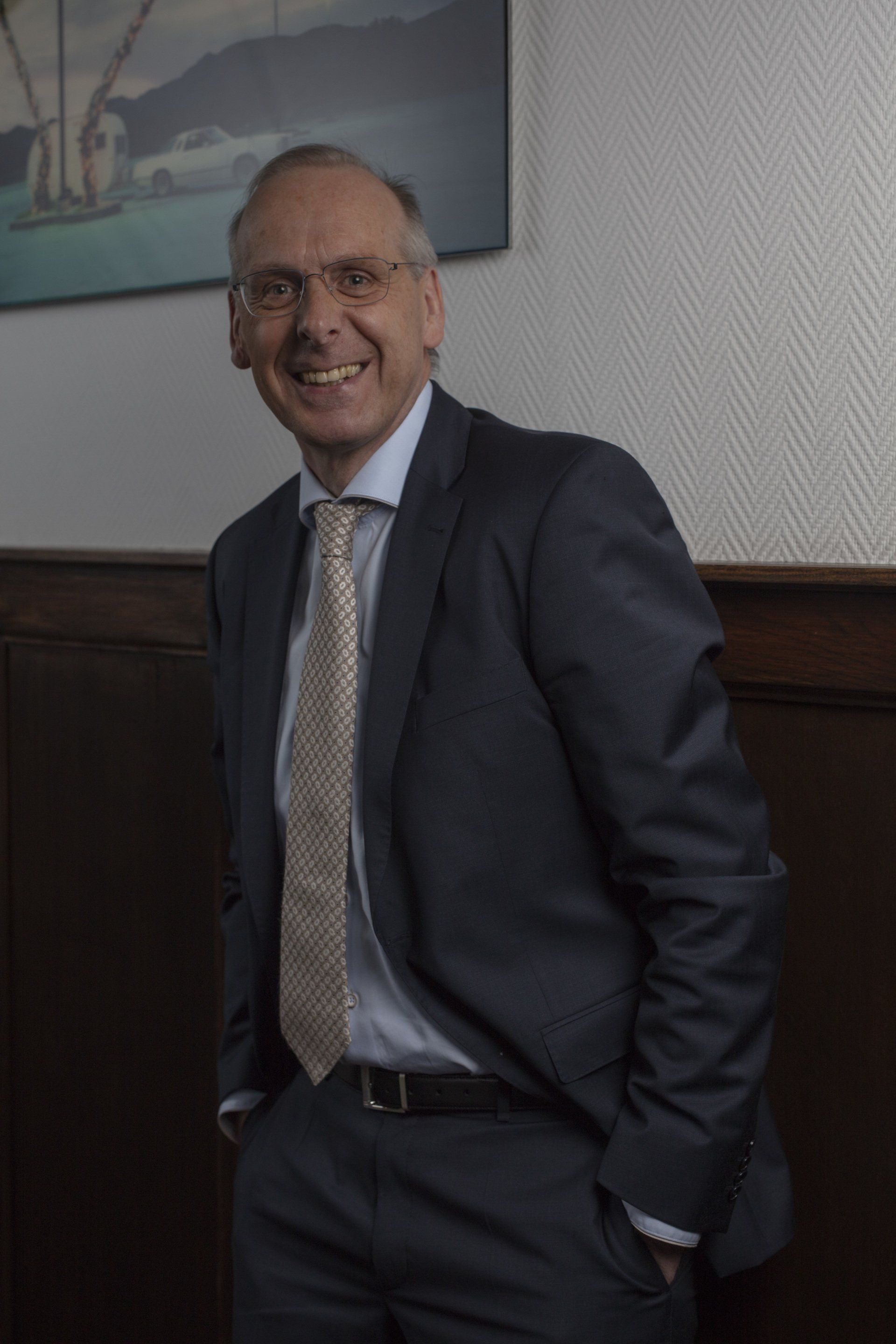 Johan Reinhoudt, glimlachend tegen een muur geleund. Hij heeft een bril, een stropdas en zijn handen in zijn zakken, in een kamer met een schilderij.
