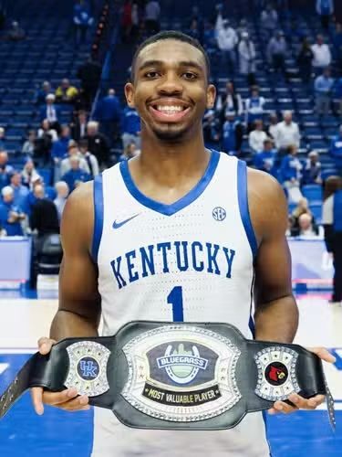 Basketball player in Kentucky jersey holds championship belt, smiling. Blue and white court setting.