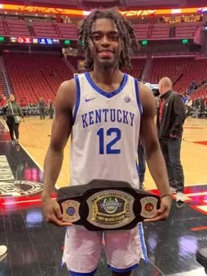 Basketball player in a Kentucky jersey, holding a championship belt on a basketball court.