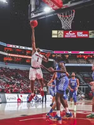 Basketball player shooting a jump shot, court with red and blue team colors, fans in the stands.