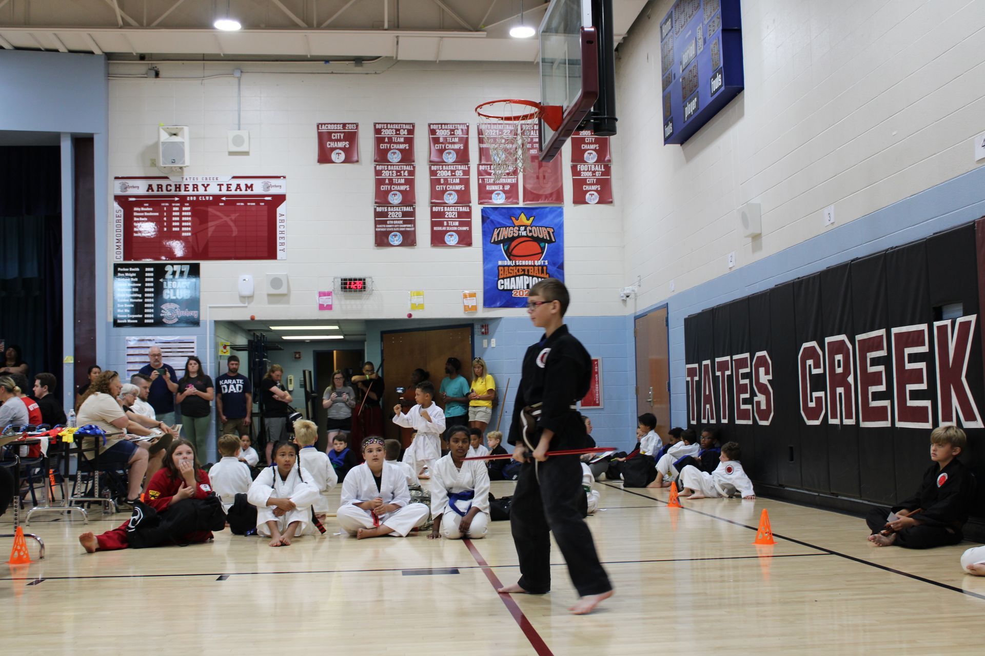 Boy in black karate uniform demonstrating in a gym with spectators, cones, and banners.