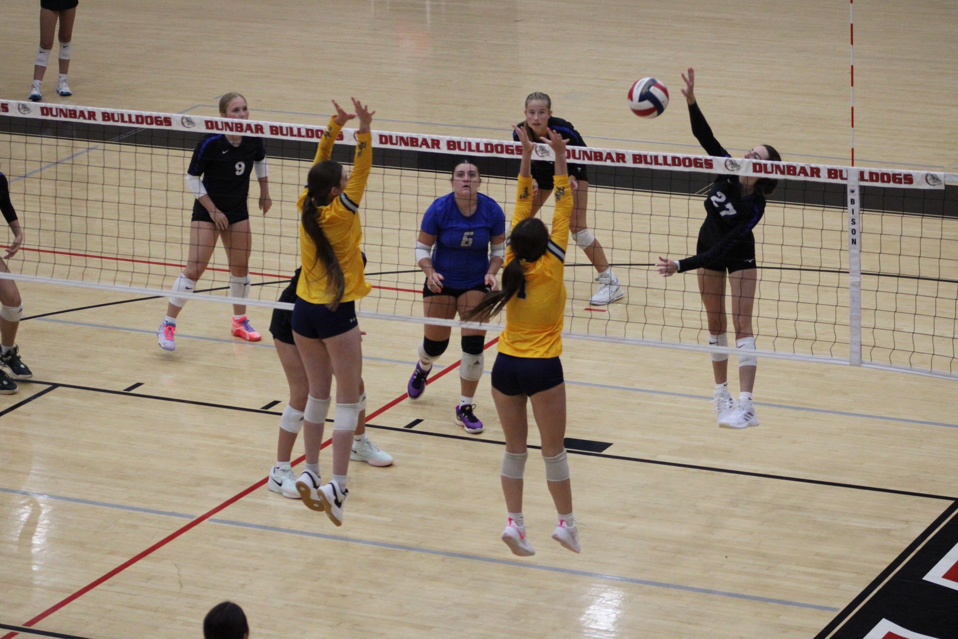 Volleyball players jumping to hit a ball over the net, in a gymnasium. Yellow and black uniforms.