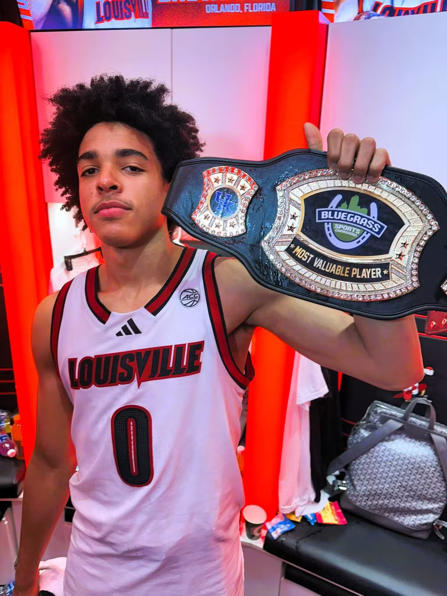 Young athlete in a Louisville jersey holds a championship belt, smiling in a red-lit room.