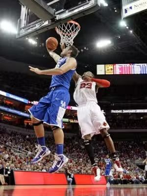 Basketball player in blue jersey attempting a shot over a defender in a red jersey. Game in arena.