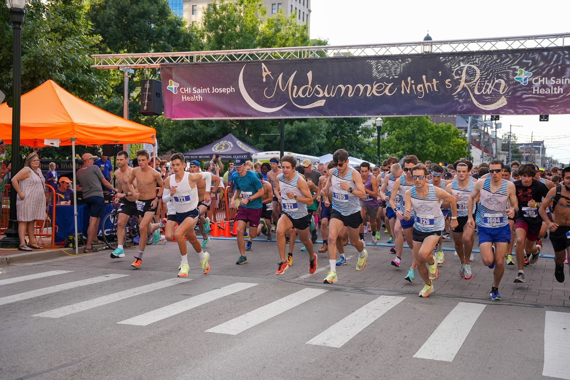Group of runners in purple shirts and race bibs pose outside near a tree and building.