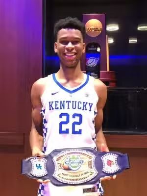 Basketball player in a Kentucky jersey holds a championship belt, smiling in front of a trophy case.