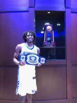 Basketball player in a Kentucky jersey holds a championship belt, trophy in background.