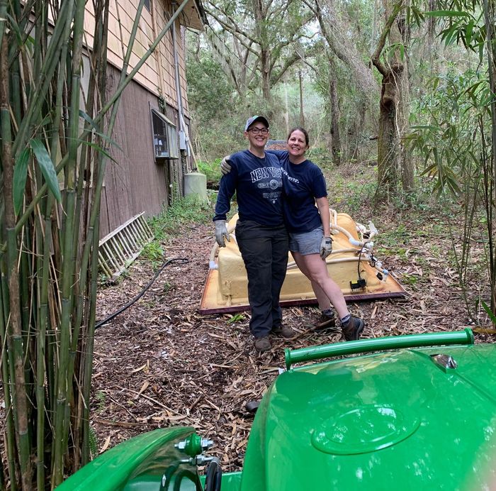 A man and a woman are standing next to a green tractor in the woods.
