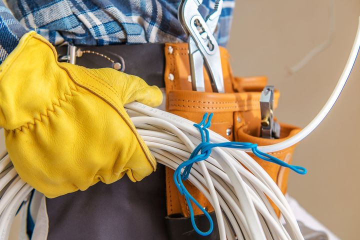 A person wearing a blue plaid shirt and yellow gloves holds a coil of white wire, with tools in a leather belt nearby.