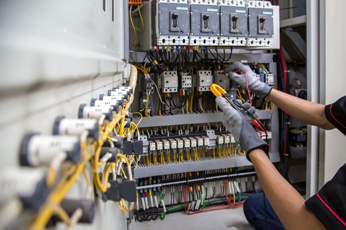 An electrician wearing gloves uses a multimeter to test electrical components inside a control panel.