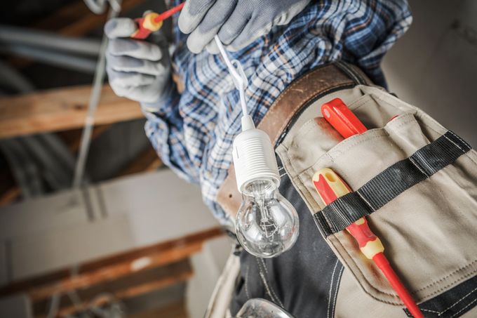 A person in work gloves holding a screwdriver and a lightbulb, wearing a tool belt with equipment at a construction site.