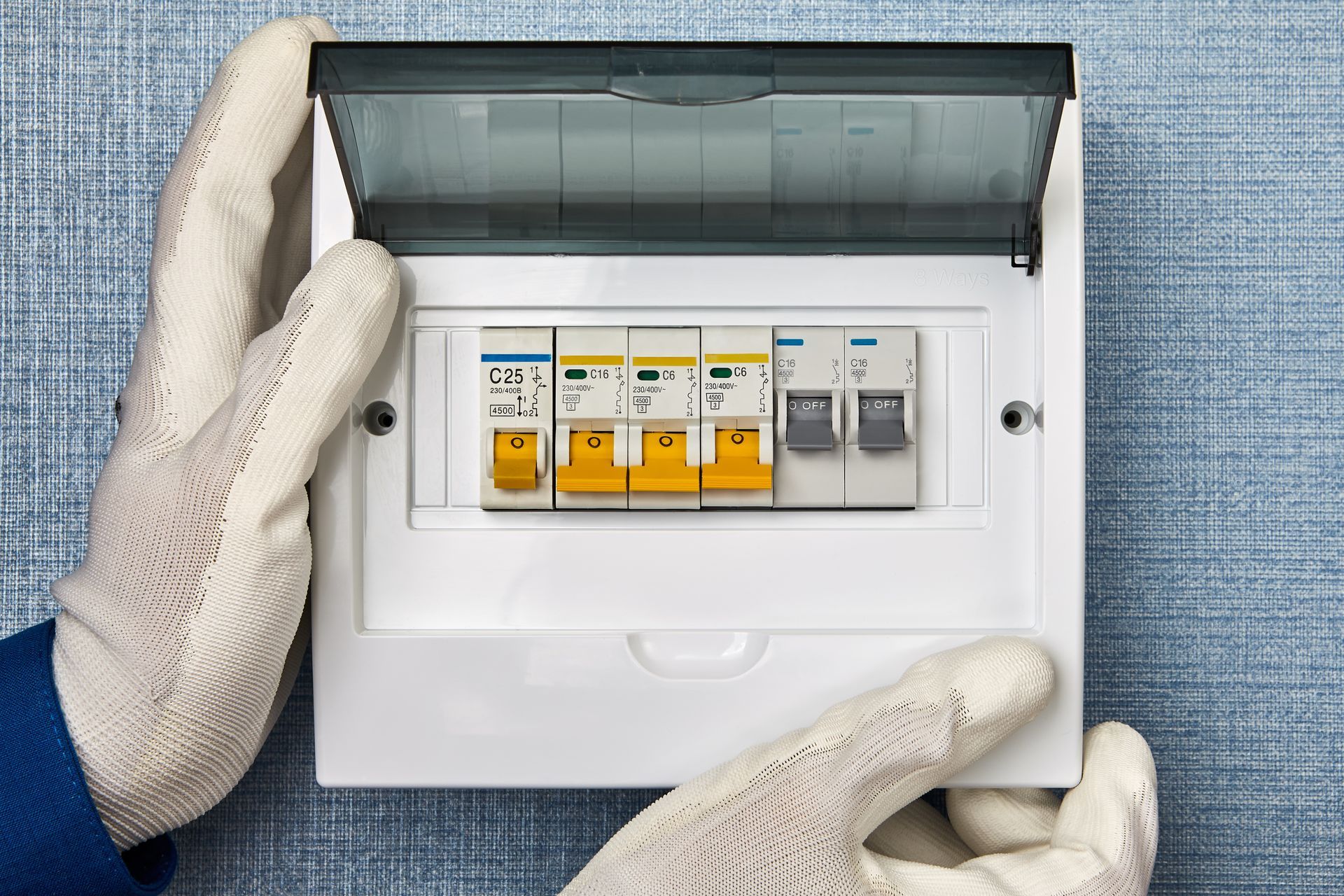 Hands in white work gloves holding an open residential electrical distribution board with several circuit breakers.
