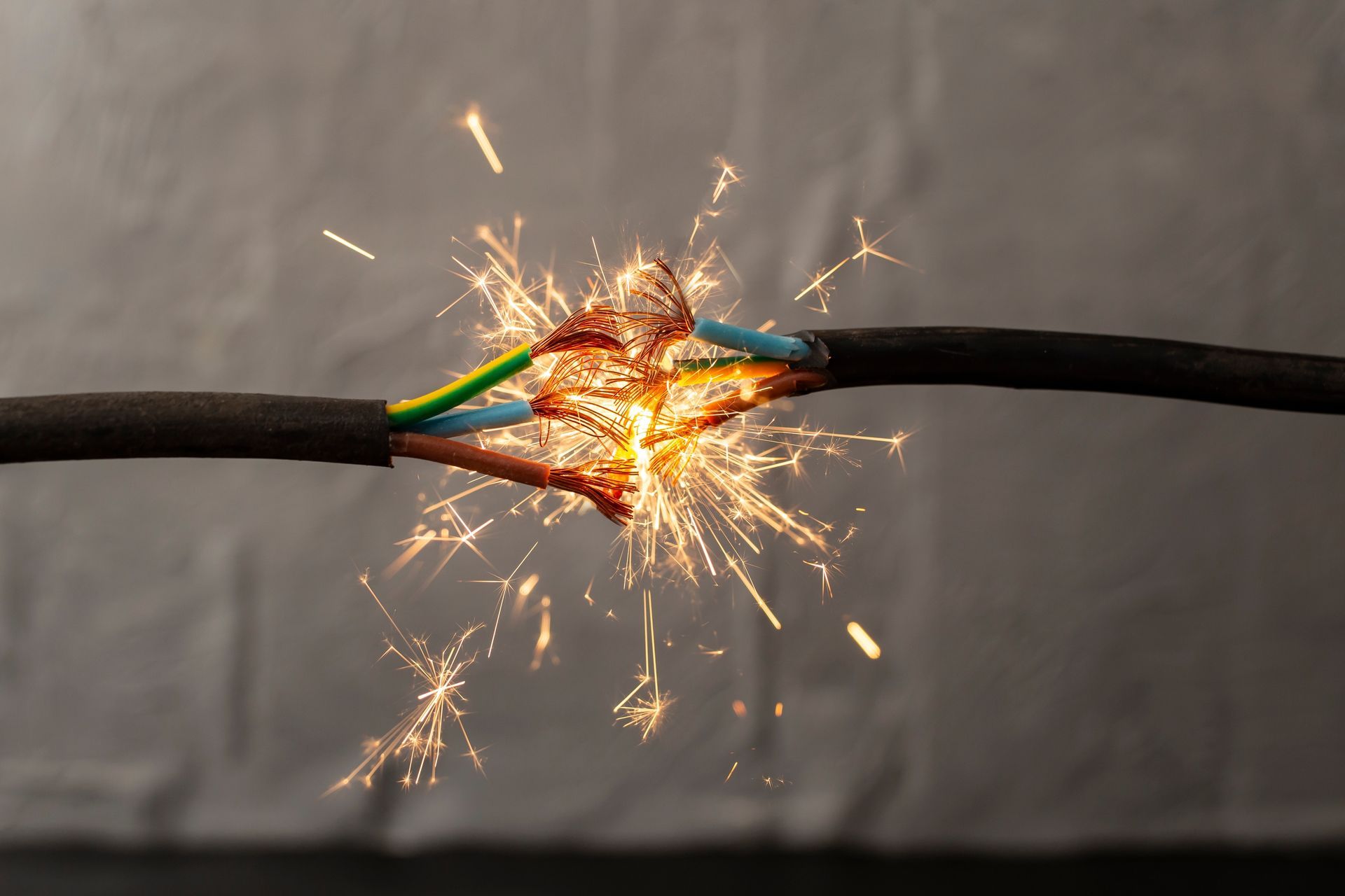 A frayed black electrical cable sparking at the point of damage against a grey background.
