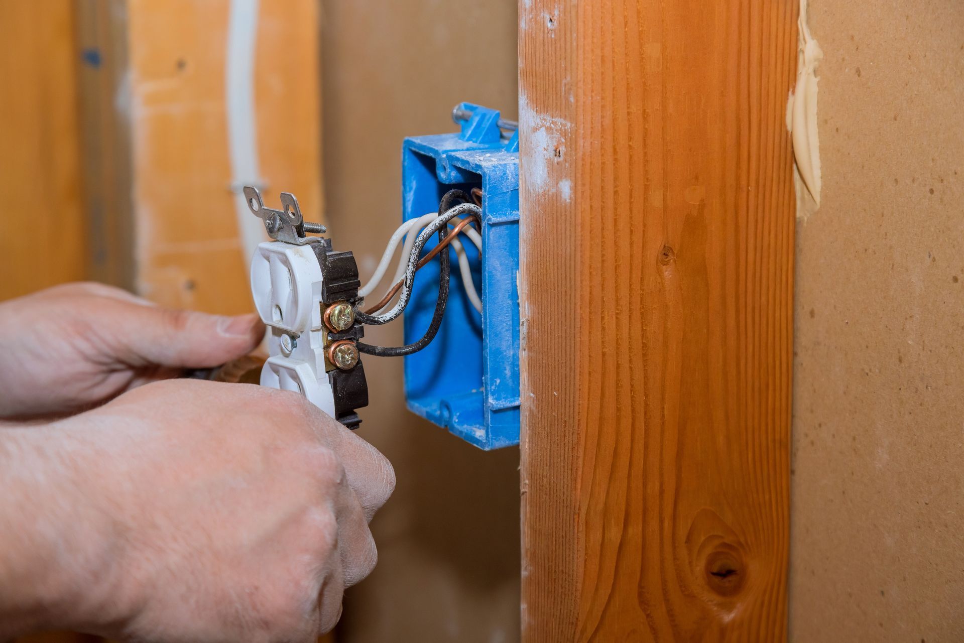 Hands wiring a white electrical outlet into a blue plastic junction box mounted on a wooden wall stud.