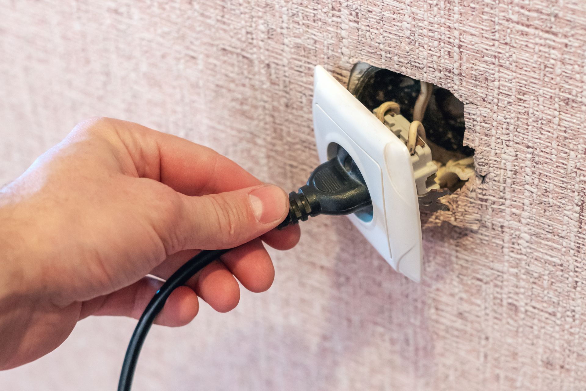 A person’s hand holds a black power cord about to plug into a loose, damaged wall electrical outlet.