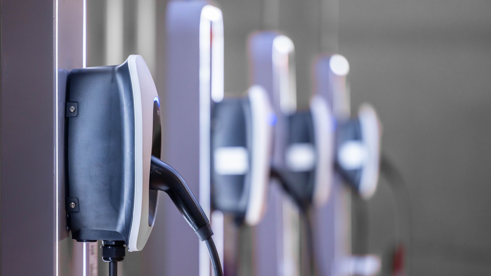 A row of wall-mounted electric vehicle charging stations with black cables plugged in, shown in a clean, modern setting.
