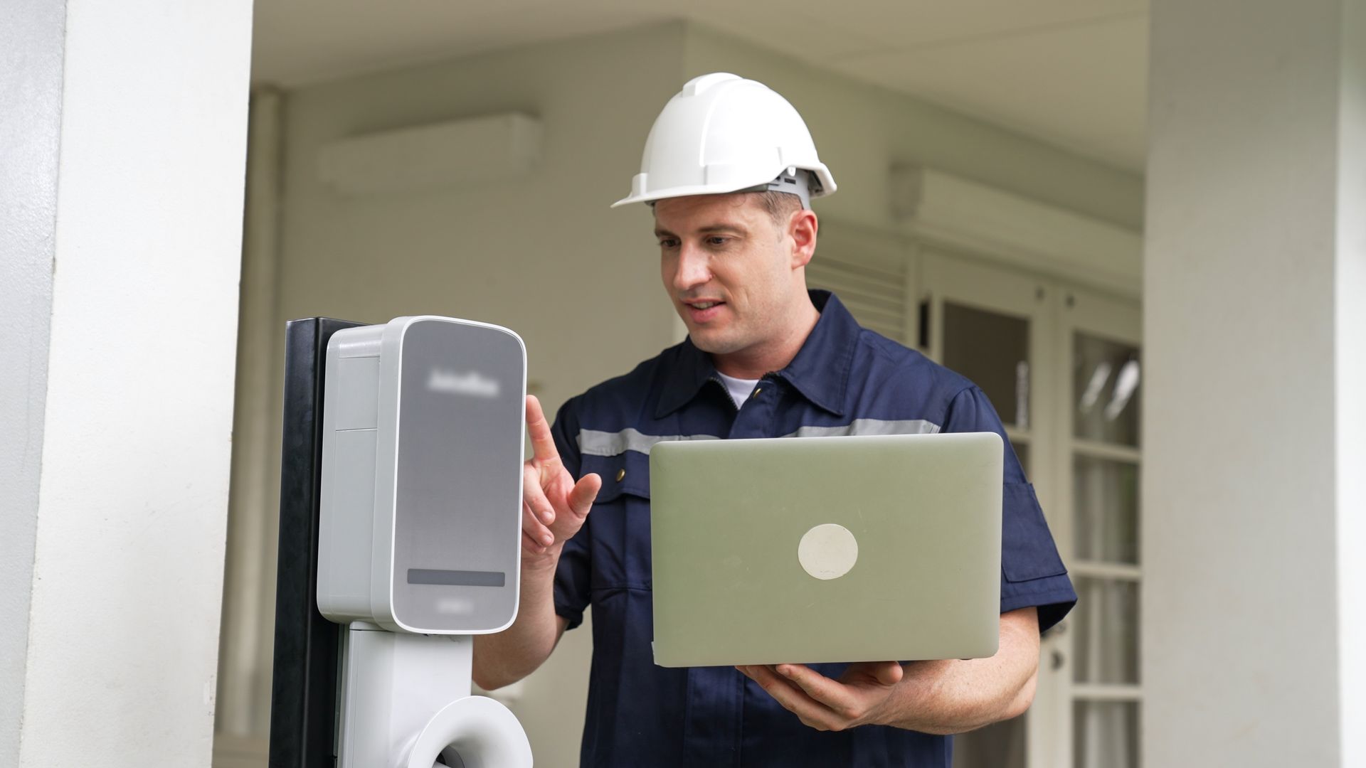 A technician in a hard hat and work uniform uses a laptop while inspecting an EV charging station.