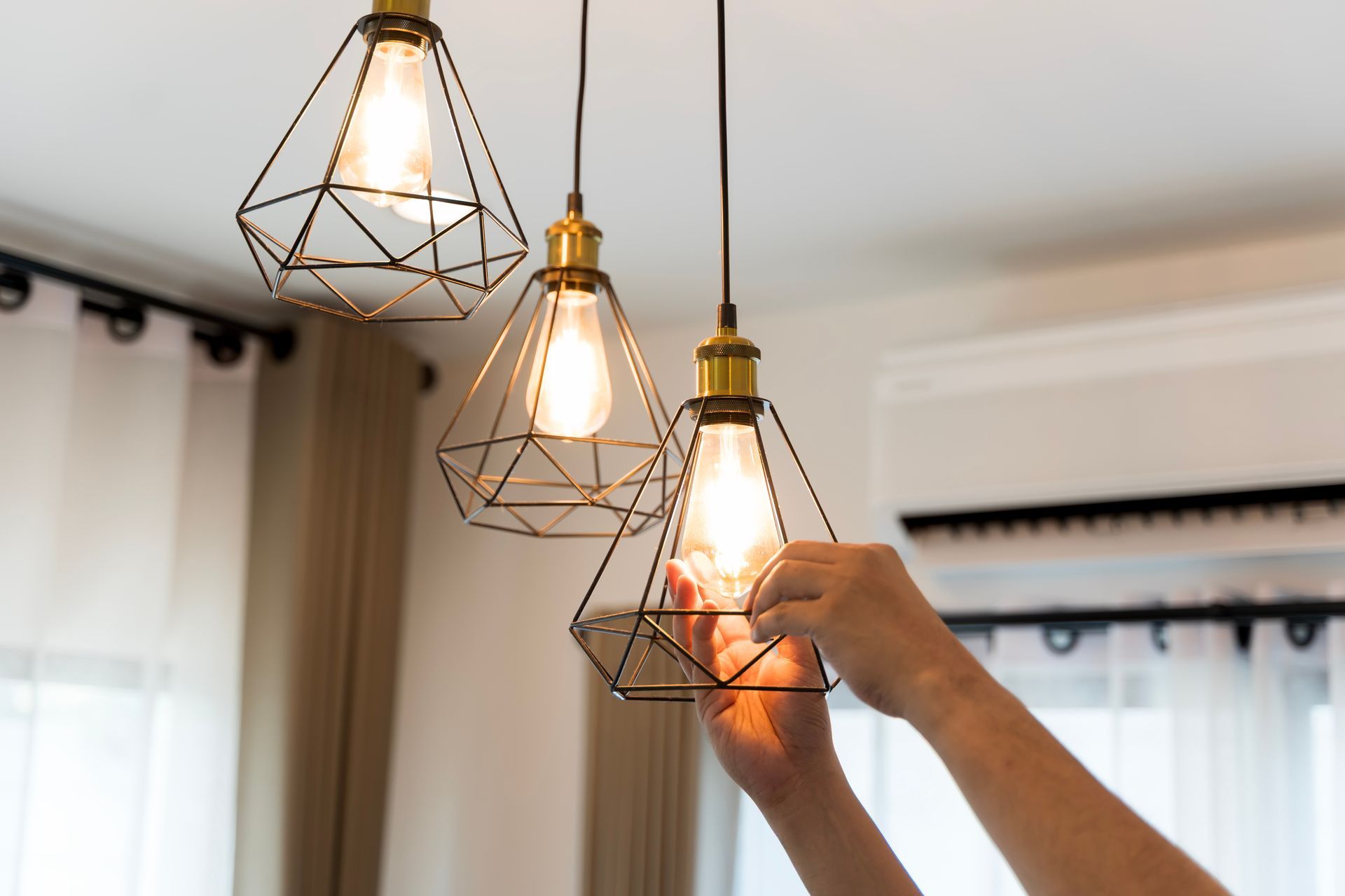 Hands adjusting a lit geometric pendant light in a room with white curtains and an air conditioner.