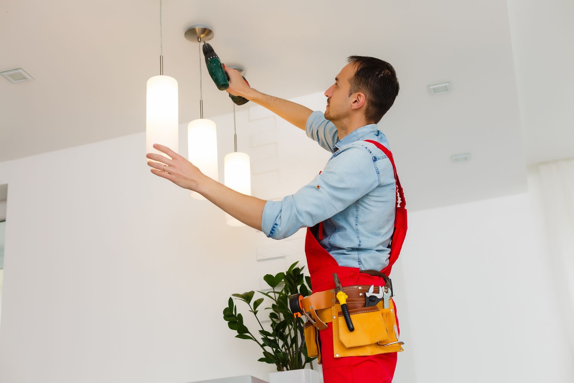 A person in red work overalls and a blue shirt uses a drill to install modern pendant lights on a white ceiling.