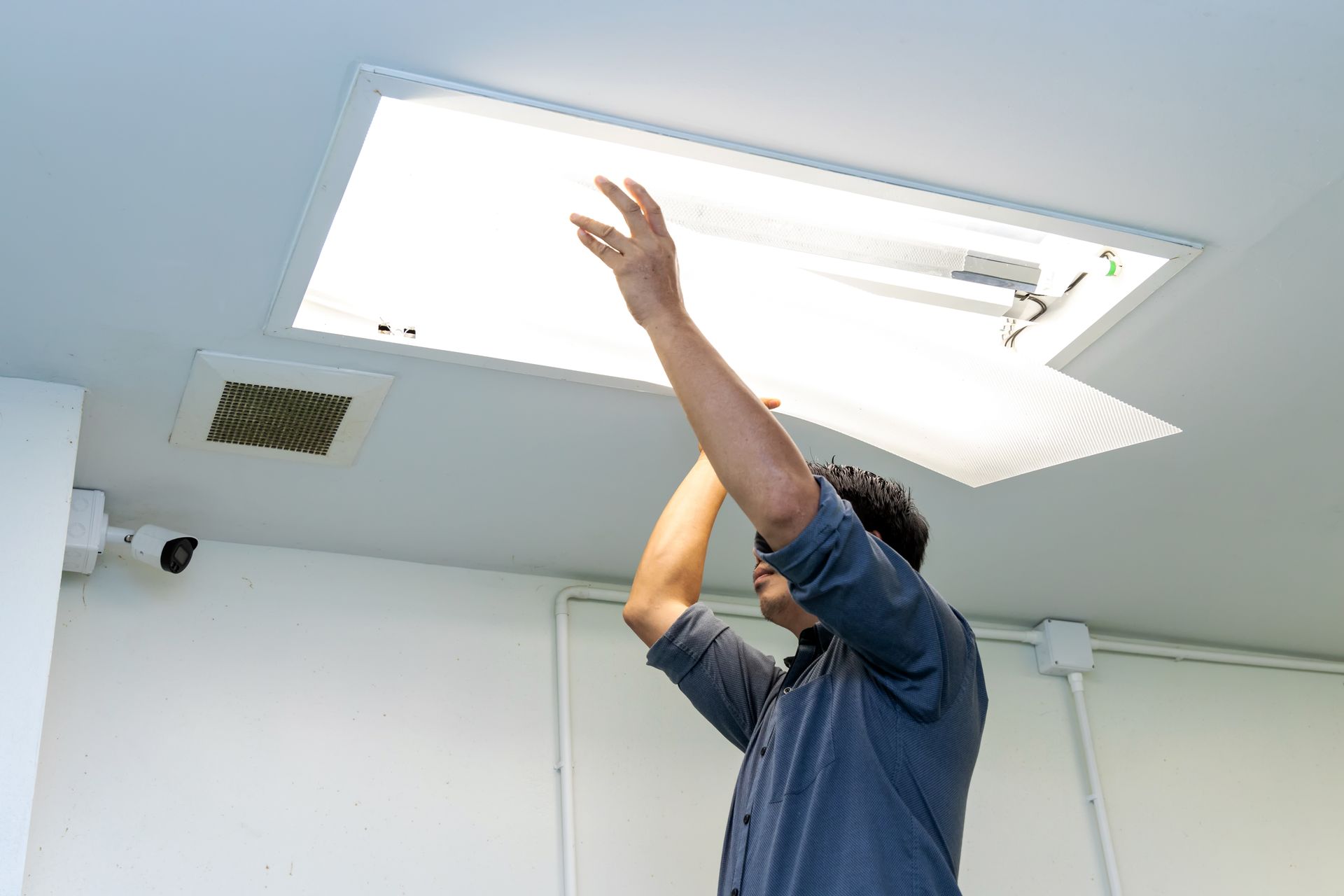 A person in a blue shirt standing on a ladder, installing or repairing a rectangular ceiling light fixture.
