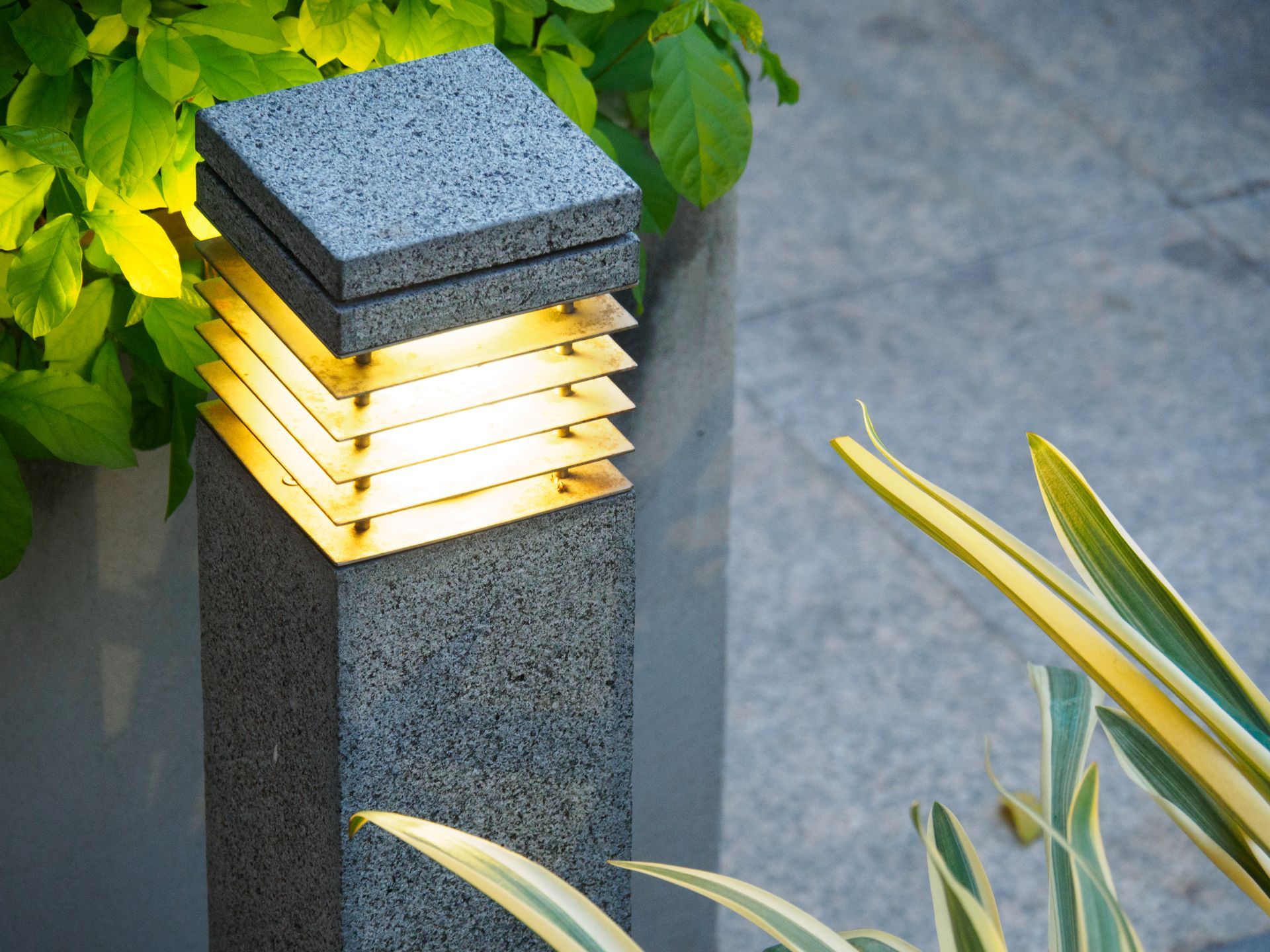 A square outdoor light post made of speckled grey stone with horizontal golden slats, situated next to greenery.